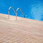 Una piscina con acqua blu e limpida. Sul bordo della piscina c'è un decking in teak. Al centro della piattaforma, c'è una scala in metallo con due corrimano curvi che scendono nell'acqua