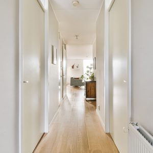 Un lungo e luminoso ma stretto corridoio di un grande appartamento, con porte da entrambi i lati, pareti chiare, pavimenti a parquet e salotto che si intravede in fondo