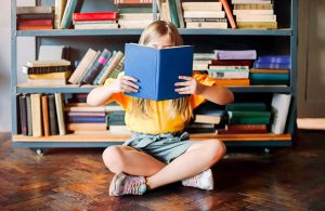 Una bambina bionda seduta a gambe incrociate su un parquet in una stanza con una libreria piena di volumi, sta leggendo un libro che le copre il volto