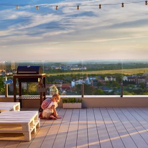 Un bambina piccola con zainetto sulle spalle, accovacciata sul decking di una grande terrazza con arredi realizzati con bancali e affacciata su un panorama verde, con ringhiera trasparente in vetro