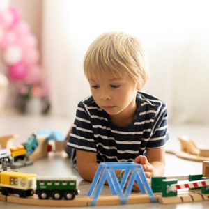 Un bambino biondo sdraiato sul parquet sta giocando con dei trenini su un binario di legno