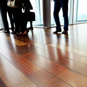 Dettaglio sul pavimento in parquet di un ufficio open space con molte finestre e persone di cui non si vedono i volti riunite a parlare