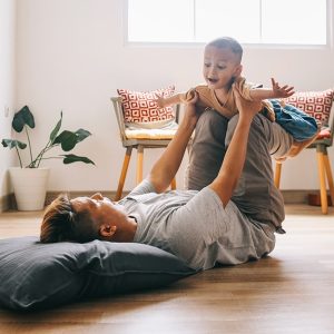 Un padre sdraiato su un pavimento in parquet, con la testa appoggiata a un cuscino sta giocando con su figlio piccolo sollevandolo in aria come fosse un aeroplano