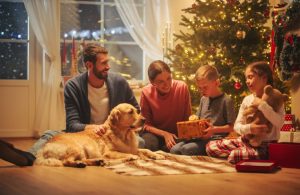 Una famiglia felice composta da padre, madre, figlia e figlio, sta seduta sul parquet a scartare regali, insieme al cane, accanto all'albero di Natale, la sera