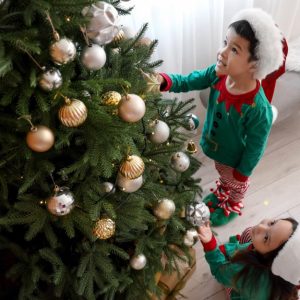 Vista dall'alto, di tre quarti, di un bambino e una bambina in pigiama e con in testa il cappello di Babbo Natale che stanno addobbando un albero di Natale con palline dorate e argentate