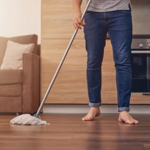 Un uomo scalzo, del quale si vede solo dal busto in giù, sta passando lo straccio sul parquet in una casa con mobili moderni in legno