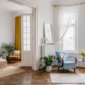 Luminoso open space con pareti e soffitto bianchi, parquet scuro, letto, poltrona, tappeto, porta a vetri e molte piante, con mobili dai colori d'accento accesi