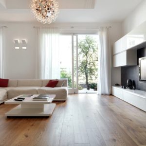 Salotto moderno in stile minimal tutto sui toni del bianco, con sofà, grande televisore, tavolino da caffè, due porte-finestra, lampadario di design e parquet a grandi plance