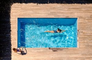 Vista dall'alto di una piscina domestica all'aperto, con una donna che fa il bagno e un decking in legno chiaro che circonda la piscina