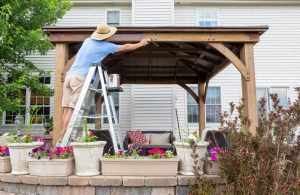 Un uomo con cappello di paglia, arrampicato su una scala, sta verniciando un gazebo in legno in un giardino con muretto di pietre e molte piante