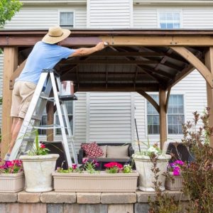 Un uomo con cappello di paglia, arrampicato su una scala, sta verniciando un gazebo in legno in un giardino con muretto di pietre e molte piante