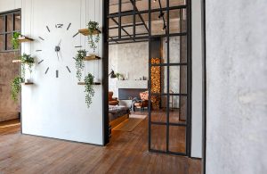 Ingresso di una casa in stile industrial minimale, con tramezzo bianco e porte a vetri nere, grande orologio sulla parete e piccole mensole con piantine. Si intravede un'altro ambiente: salotto con pareti in cemento grezzo, camino, poltrone e legna da ardere impilata. Il pavimento è in parquet scuro