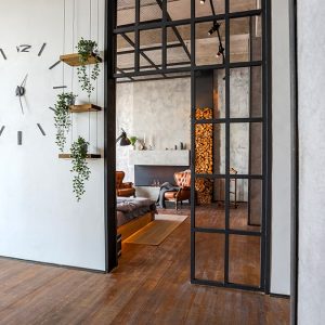 Ingresso di una casa in stile industrial minimale, con tramezzo bianco e porte a vetri nere, grande orologio sulla parete e piccole mensole con piantine. Si intravede un'altro ambiente: salotto con pareti in cemento grezzo, camino, poltrone e legna da ardere impilata. Il pavimento è in parquet scuro