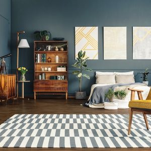 Grande camera da letto con pareti blu-grigio, pavimento in parquet scuro, letto matrimoniale sui toni del bianco e del blu polvere, poltrona in pelle giallo senape e struttura in legno, mobili vintage in legno scuro, tappeto con motivo geometrico bianco e grigio